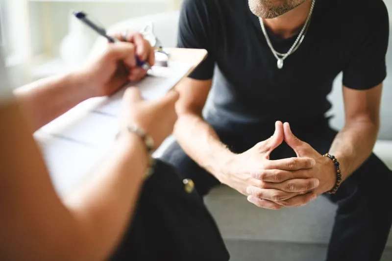 A clinician with a clipboard talks with a seated patient during a private outpatient medication-assisted treatment visit.