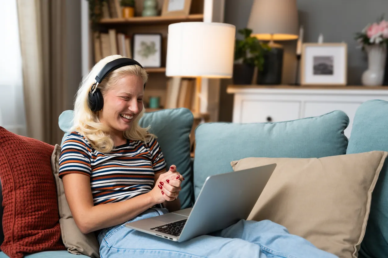A young woman sits on a sofa at home with headphones on, attending a video appointment on her laptop.