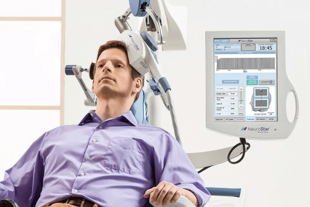 A seated patient reclines in a NeuroStar TMS treatment chair while the magnetic coil arm rests against the left side of his head during a session.