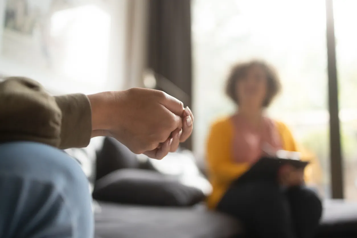 A client's clasped hands in the foreground while a therapist takes notes across the room during a one-on-one session.